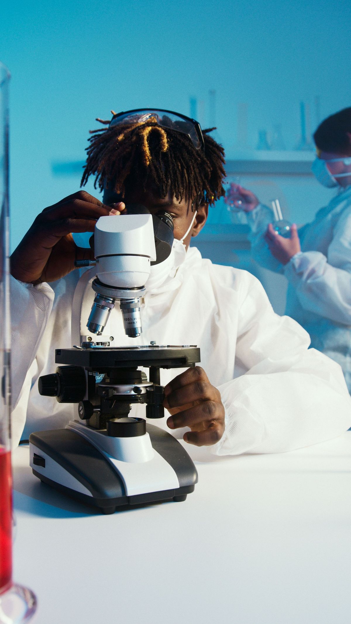 A laboratory professional using a microscope while a colleague works in the background.